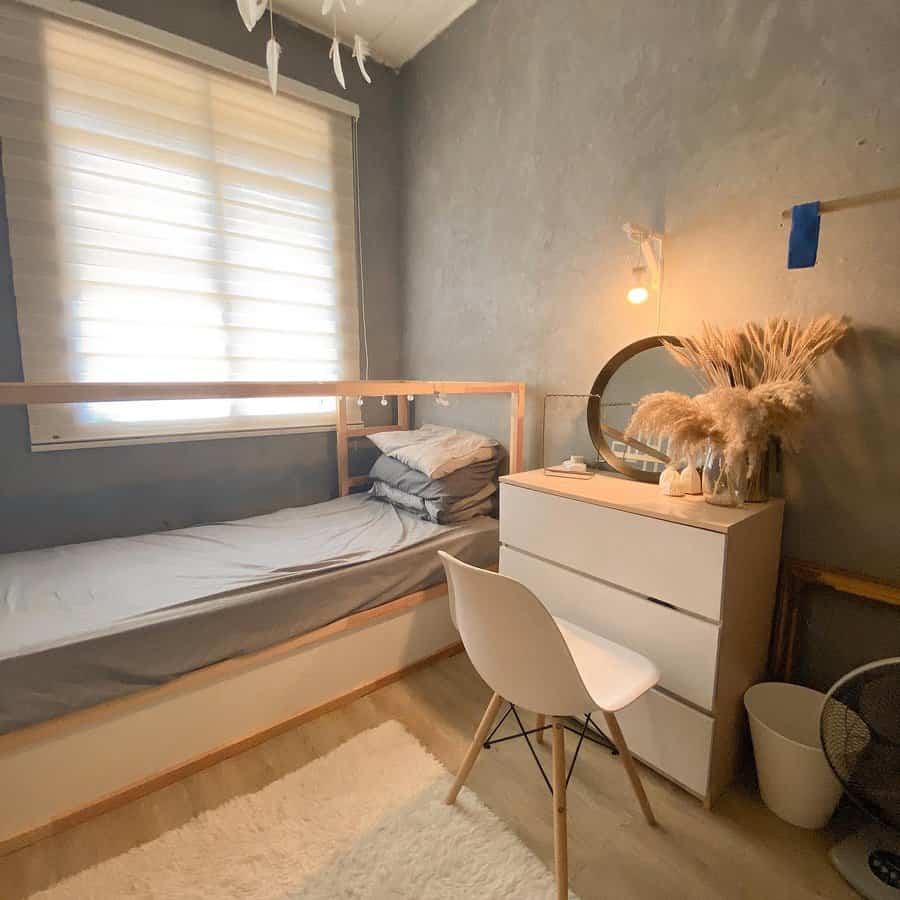 Minimalist bedroom with a single bed, white dresser, chair, round mirror, and decorative pampas grass; warm lighting and a cozy rug