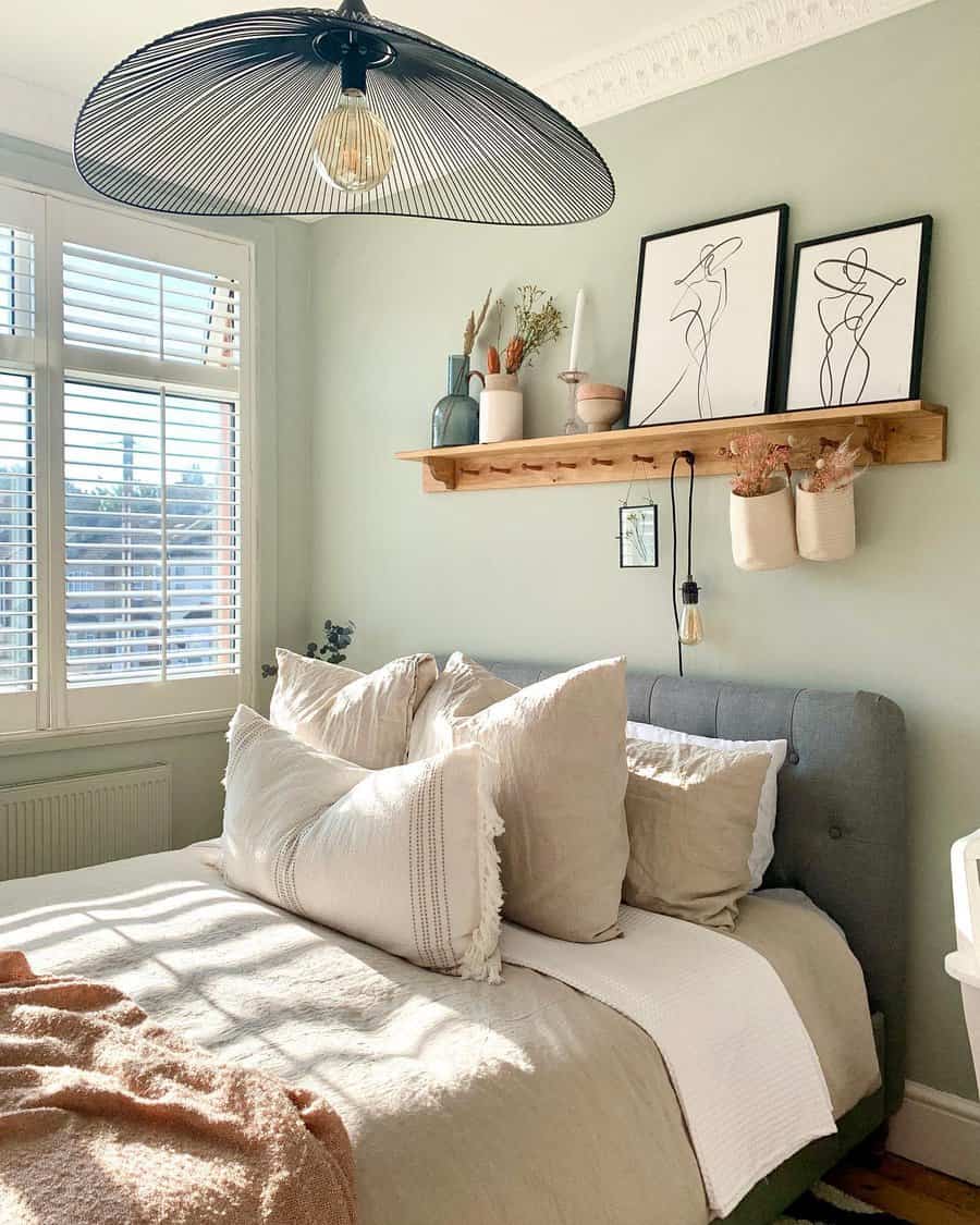 Cozy bedroom with a gray upholstered bed, fluffy pillows, and a wooden shelf with decor and art, large window with shutters