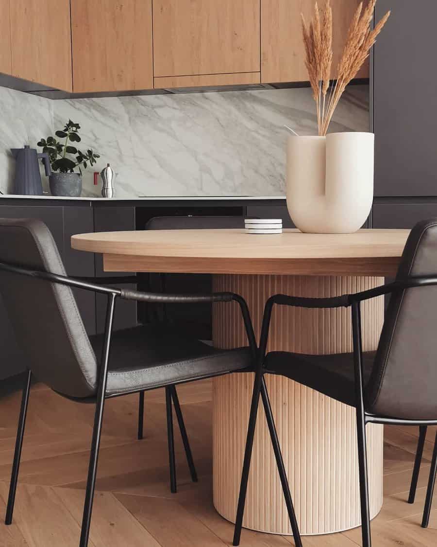 Modern kitchen with a round wooden table, two black chairs, a white vase with dried plants, and minimalist decor