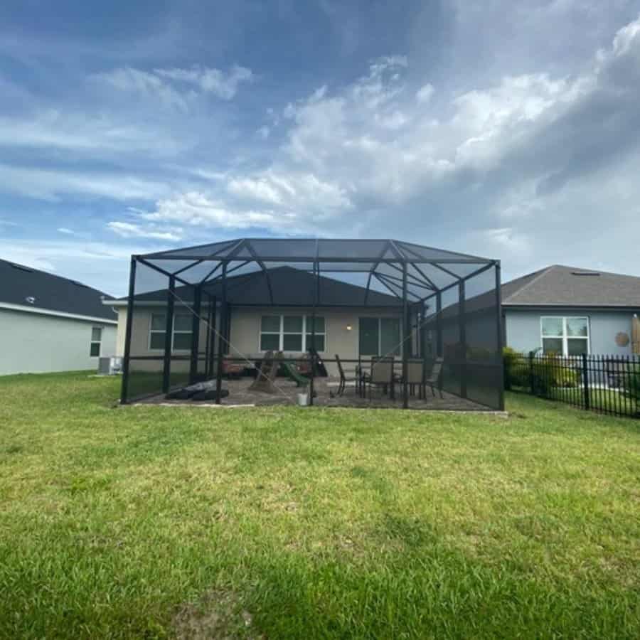 A screened patio enclosure with patio furniture in a backyard, set against a cloudy sky and surrounded by grass and neighboring houses