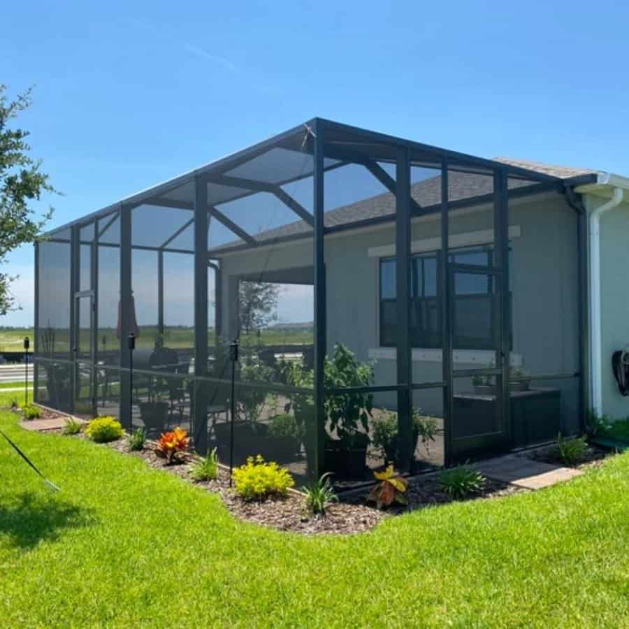 Screened-in patio attached to a house, surrounded by green grass and plants under a clear blue sky