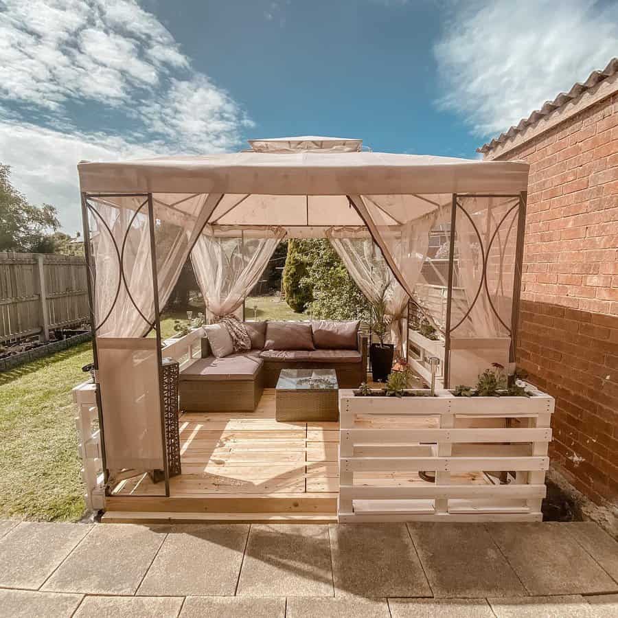 Outdoor gazebo with sheer curtains, patio furniture, and plants on a wooden deck under a sunny blue sky with scattered clouds