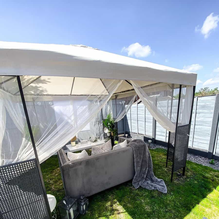 Outdoor gazebo with white canopy, sheer curtains, and a gray sofa; there's a small table and potted plant on grass under a clear blue sky