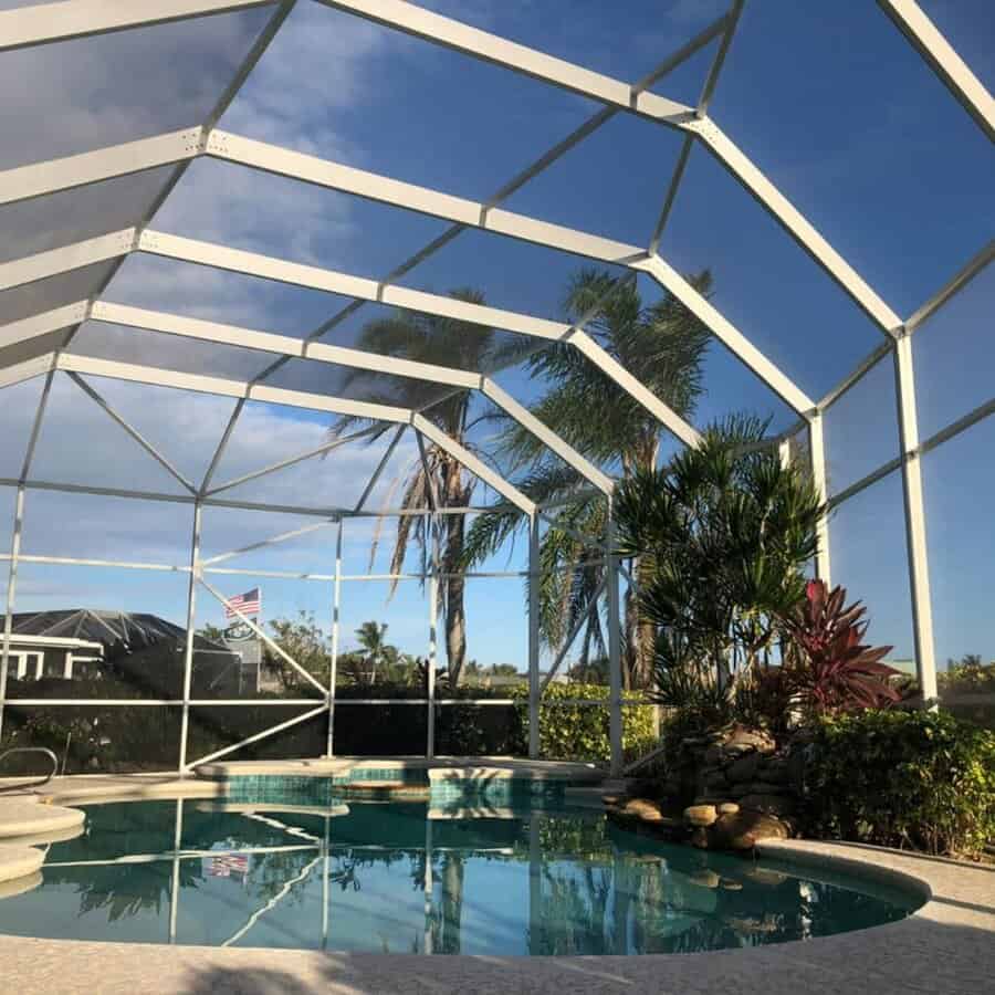 Enclosed patio with a pool, surrounded by tropical plants, clear blue sky visible through the glass ceiling and walls