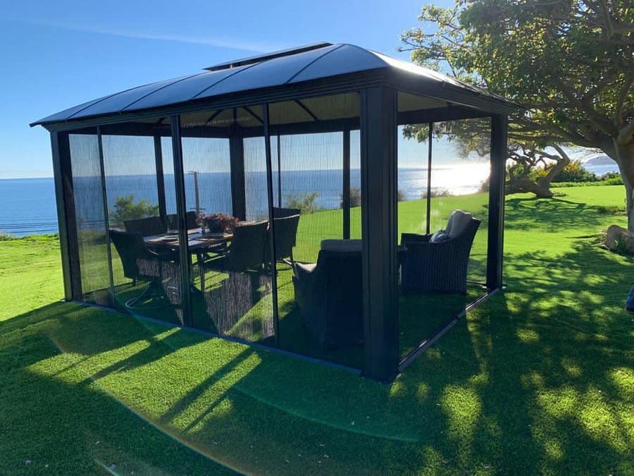 A screened gazebo with outdoor furniture is set on a grassy hill overlooking the ocean under a clear blue sky