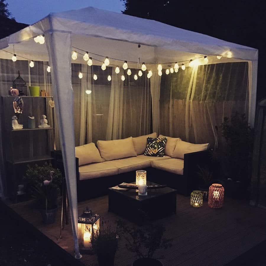 Cozy outdoor canopy with string lights, corner sofa with cushions, lanterns on the floor, and decorative shelf at night