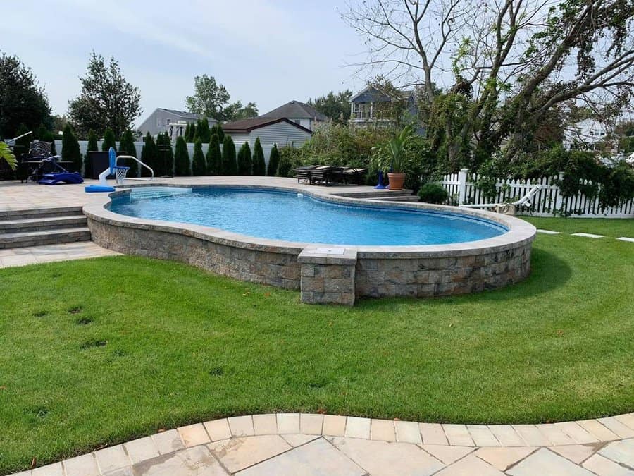 Backyard with kidney-shaped pool, stone surrounding, green lawn, and trees; houses and bushes visible in the background