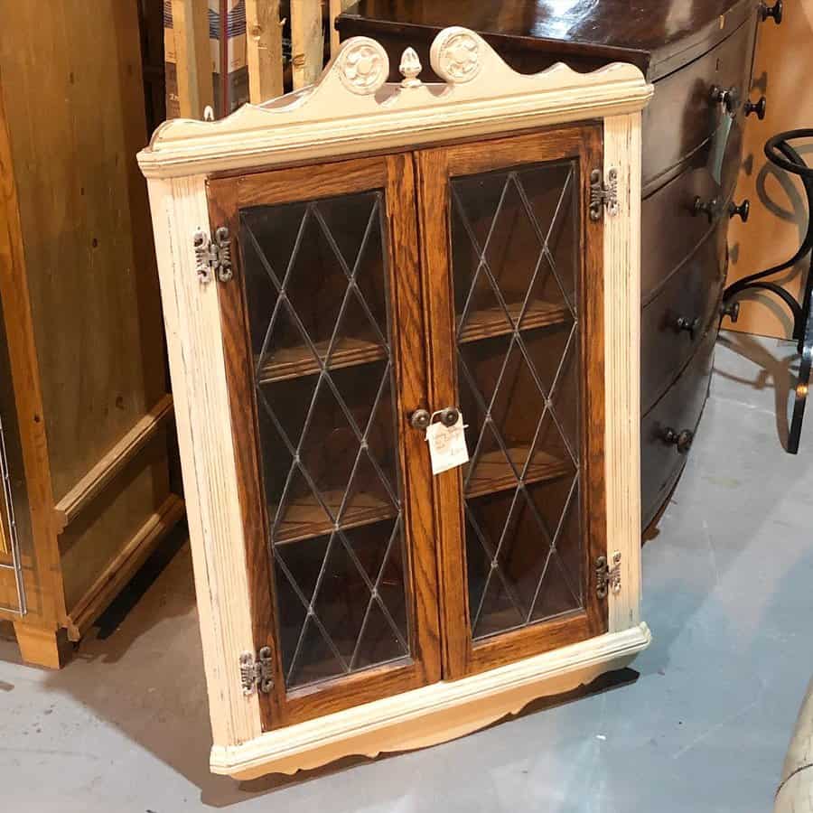 Vintage wooden cabinet with distressed cream trim and diamond-pattern glass doors, featuring ornate details and metal hinges.