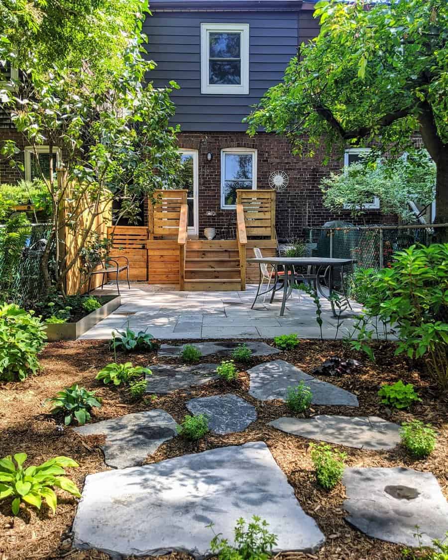 Lush garden with stone path leading to a wooden patio and stairs, surrounded by green plants and trees