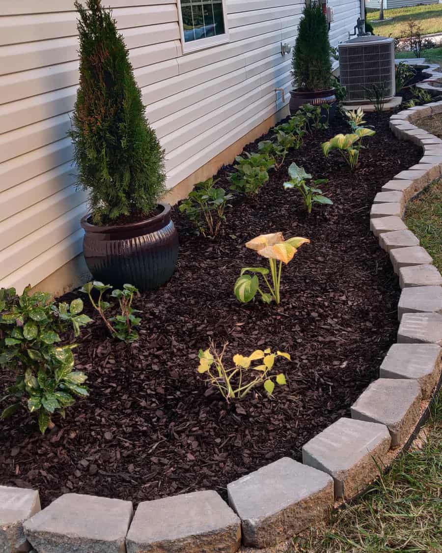 Curved garden bed with plants and mulch next to a white house, bordered by light stone edging