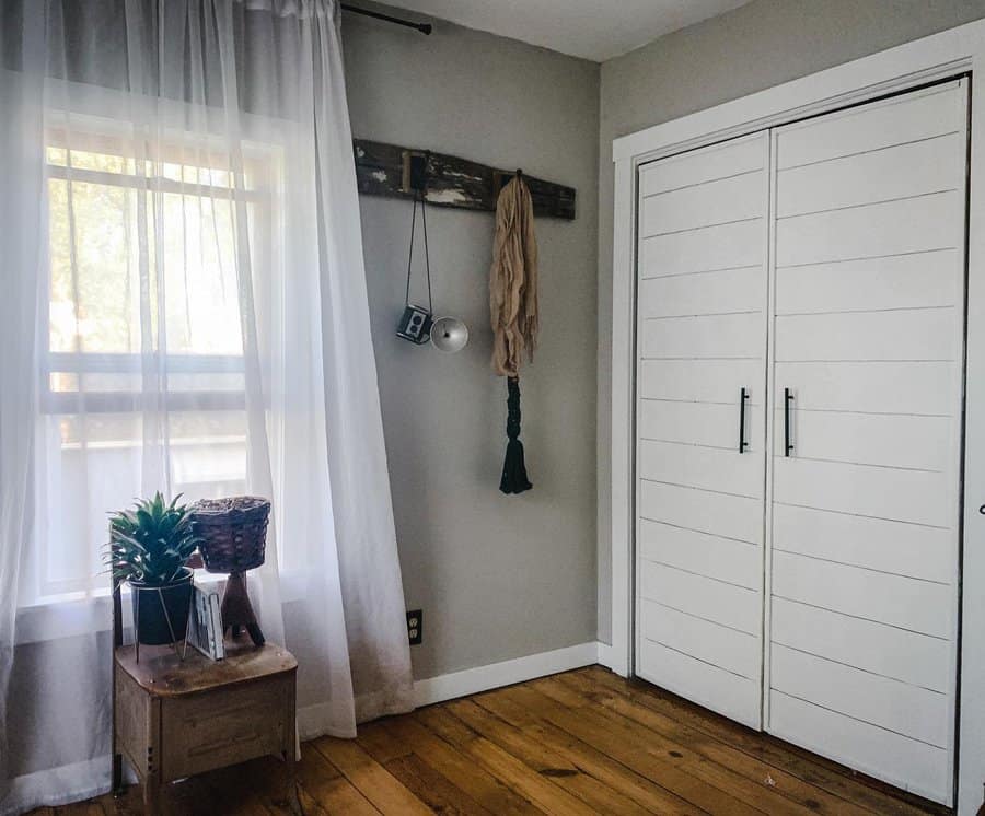 Minimalist room with white closet doors, sheer curtains, a wooden floor, and a small table with plants by the window; wall hooks hold decor