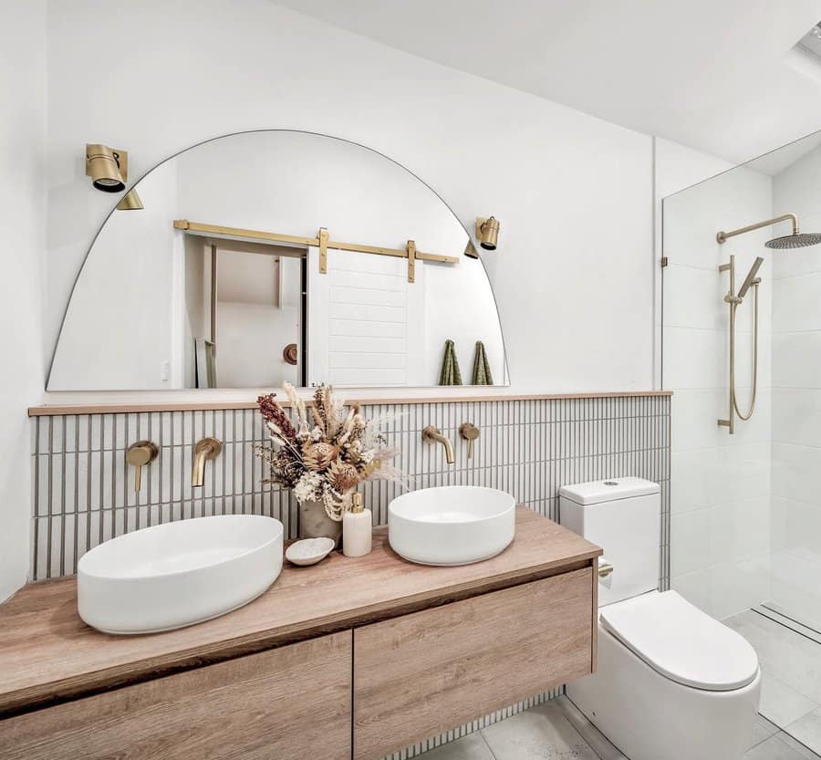 Modern bathroom with double sinks on a wooden vanity, arch mirror, and glass shower, dried flowers in vase as decoration