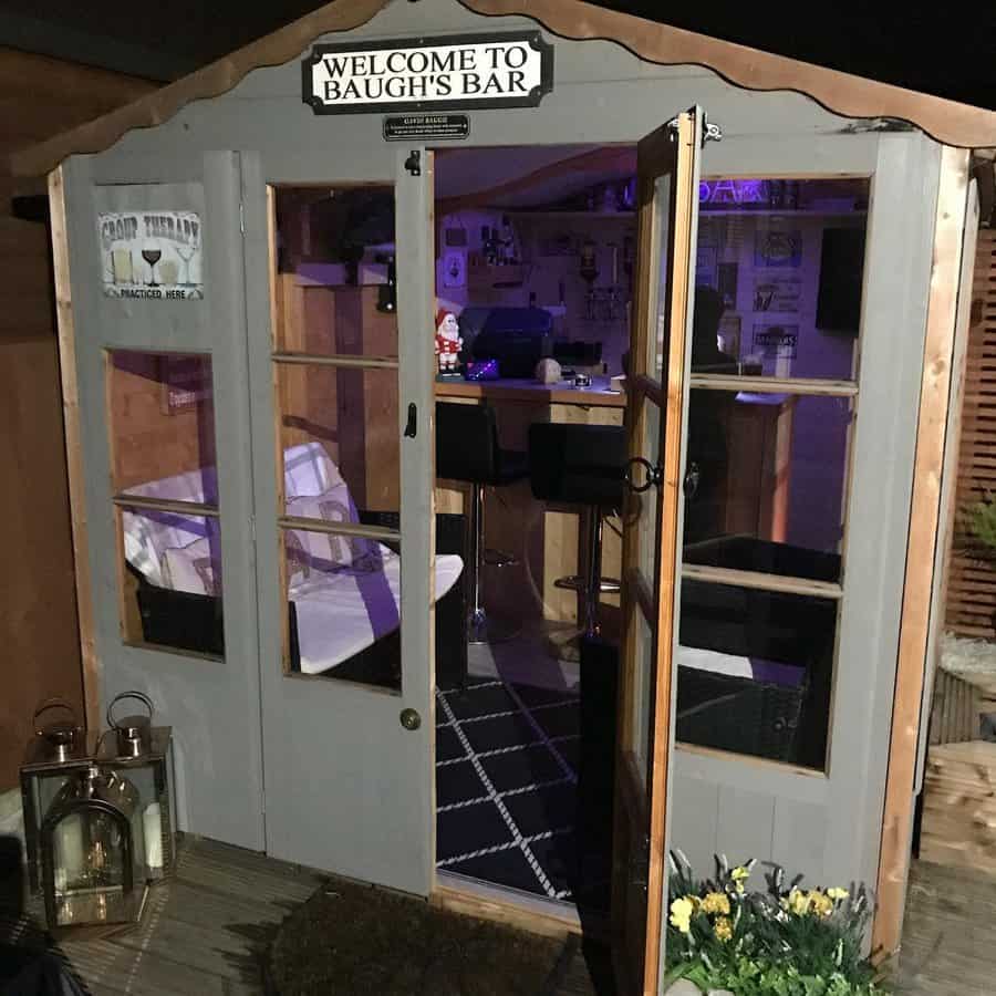 Small garden shed turned into a bar with a "Welcome to Baugh's Bar" sign, bar stools inside, and lanterns outside