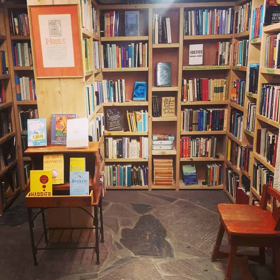 Cozy bookstore or library corner with wooden bookshelves filled with a diverse collection, a stone floor, and a small reading table with chairs.