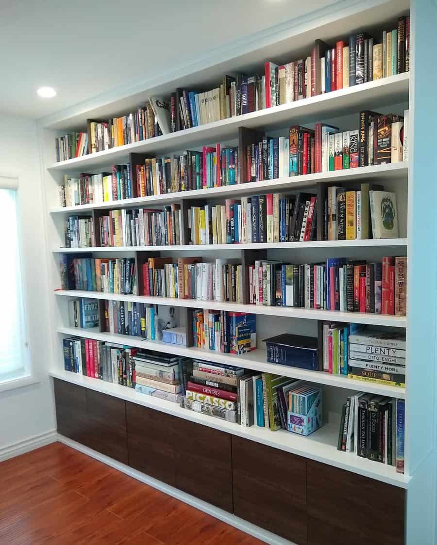 Sleek built-in white bookshelves filled with a diverse collection of books, featuring lower cabinets for extra storage in a well-lit reading space.