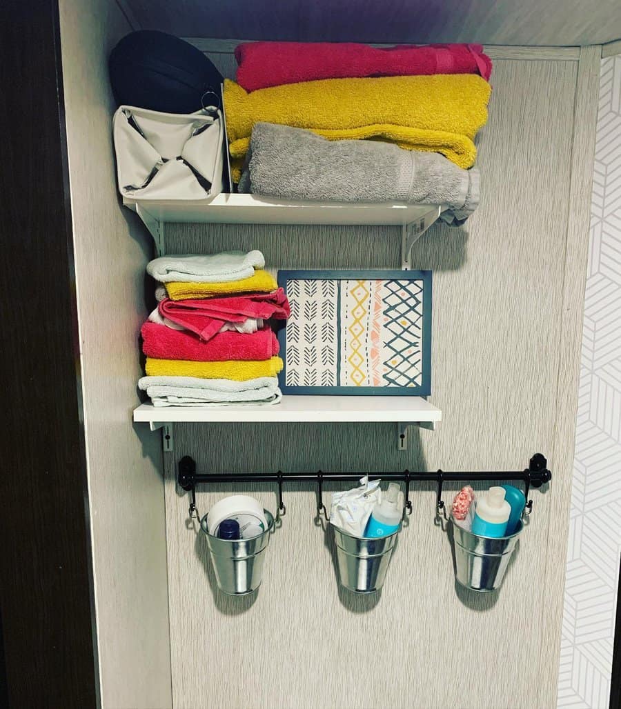 RV bathroom storage with white floating shelves for towels and decor, plus a hanging rod with metal buckets for organizing toiletries.