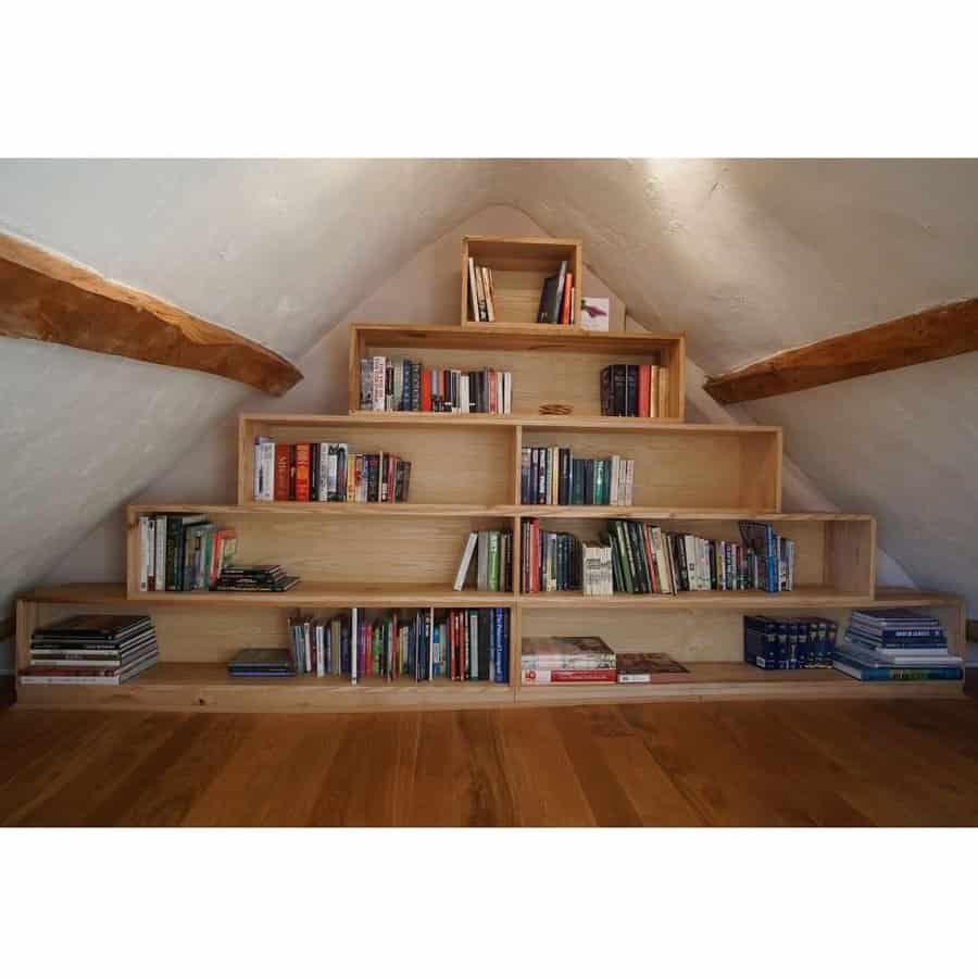 Pyramid-style wooden bookshelf in an attic with sloped ceilings, filled with books of various sizes, blending with rustic beams.