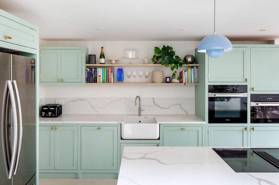 Modern kitchen with pastel green cabinets, open wooden shelves, marble countertops, and a farmhouse sink under soft pendant lighting