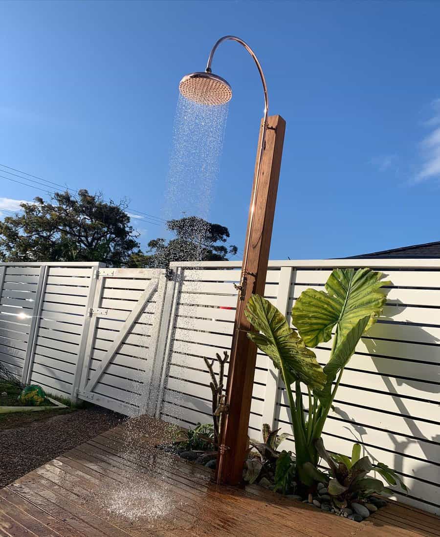 Outdoor wooden shower with running water next to a white fence, surrounded by greenery, under a clear blue sky