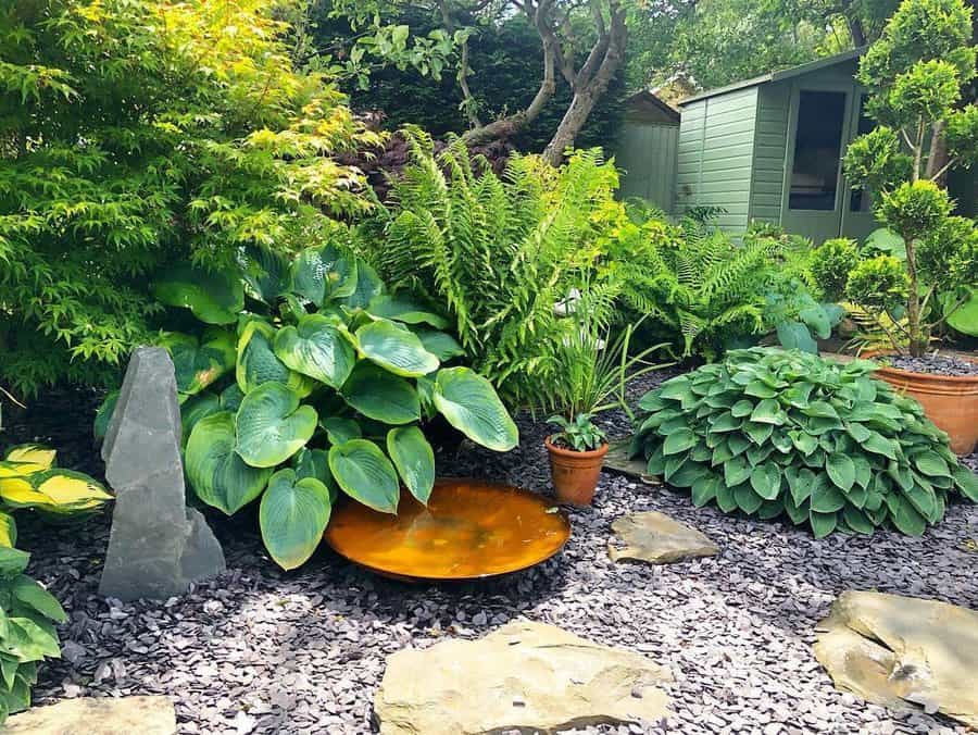 Lush garden with hostas, ferns, a small water feature, stone pathway, and a green shed in the background