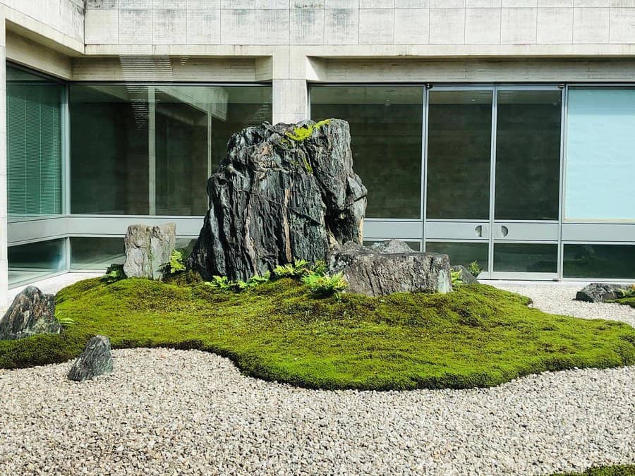 A minimalist rock garden with large stones, green moss, and gravel, set against modern concrete walls and large glass windows