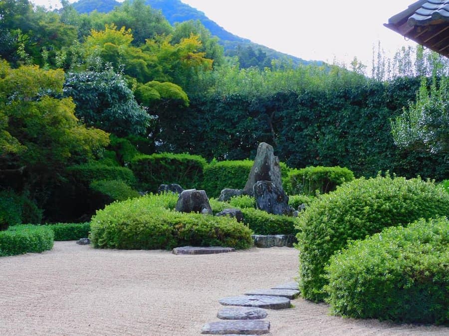 A serene Japanese garden with neatly trimmed shrubs, large stones, and a stone path, set against a backdrop of green hills