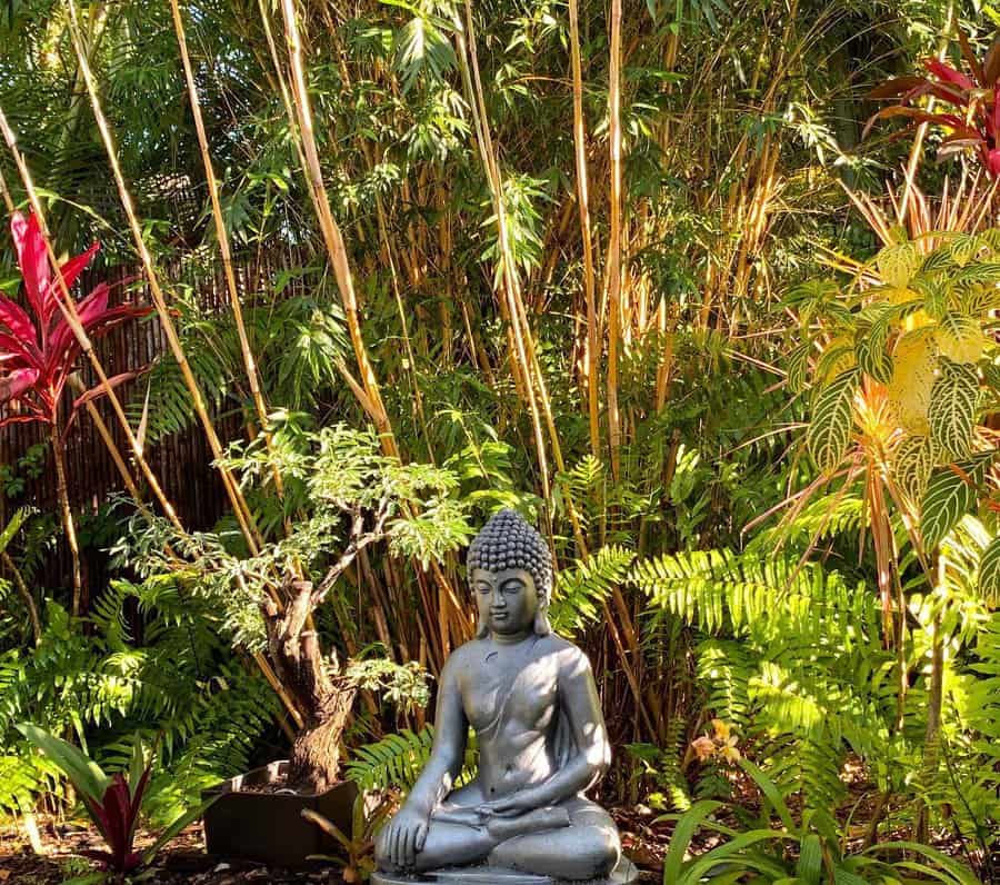 Buddha statue in a serene garden, surrounded by lush green ferns, bamboo, and vibrant red and yellow tropical plants