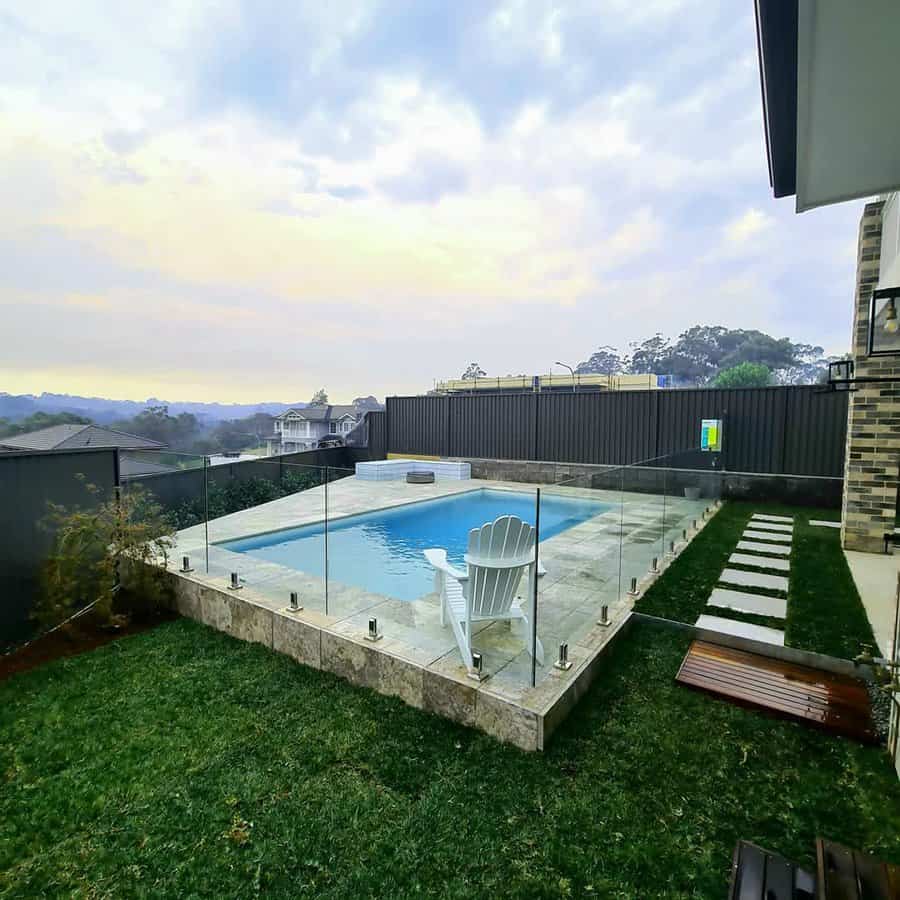 A backyard pool with a white chair beside it, surrounded by grass and a glass fence