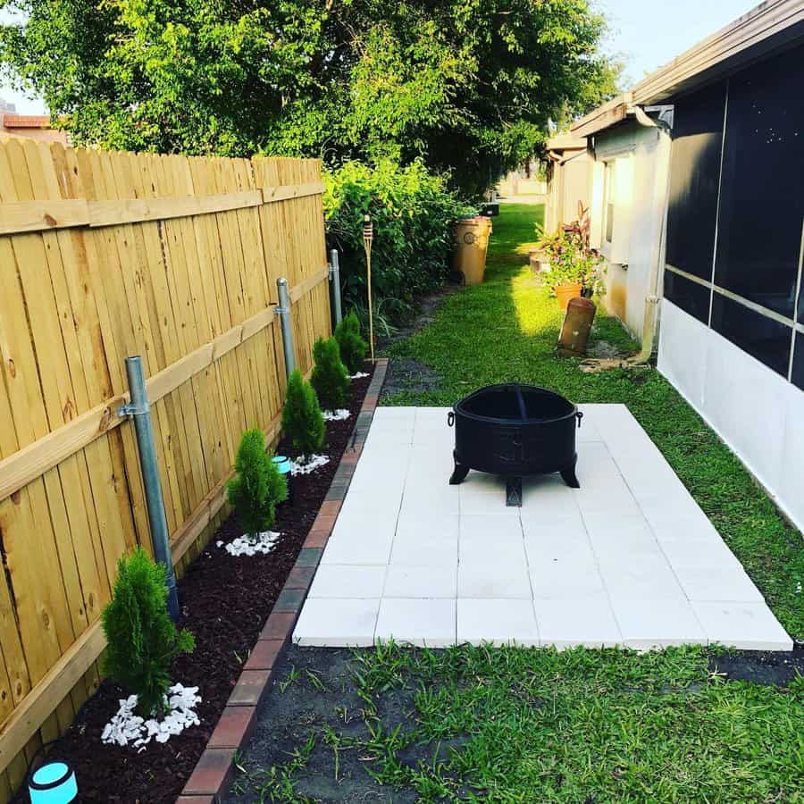 Fire pit on a tiled area with small trees along a wooden fence in a backyard