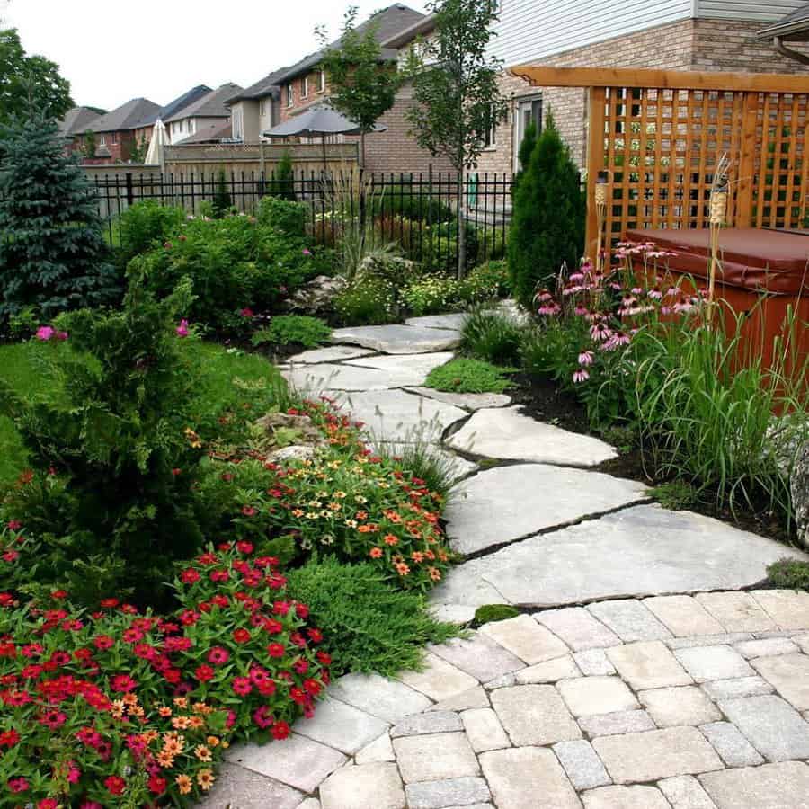 Stone path winding through a lush garden with colorful flowers, greenery, and a wooden trellis in a suburban backyard setting