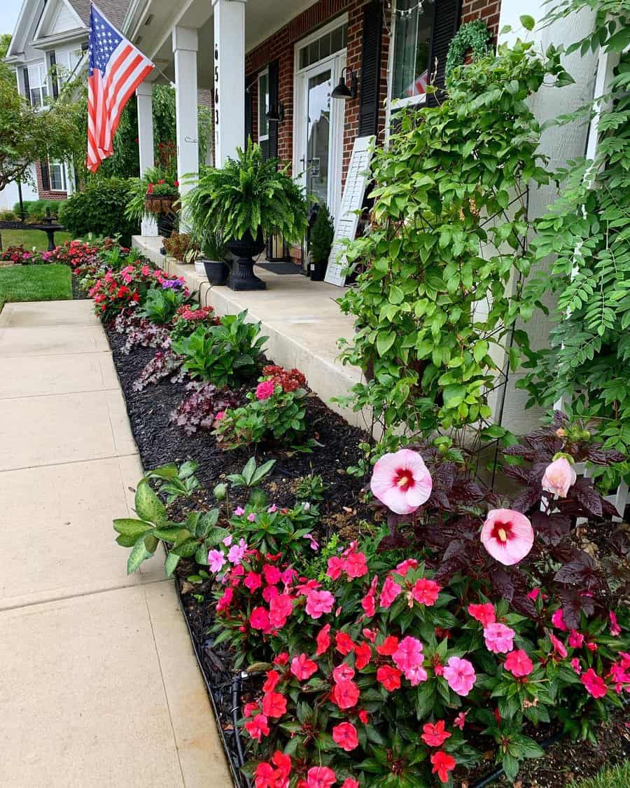 A colorful garden with pink and red flowers lines a pathway, leading to a porch with potted plants