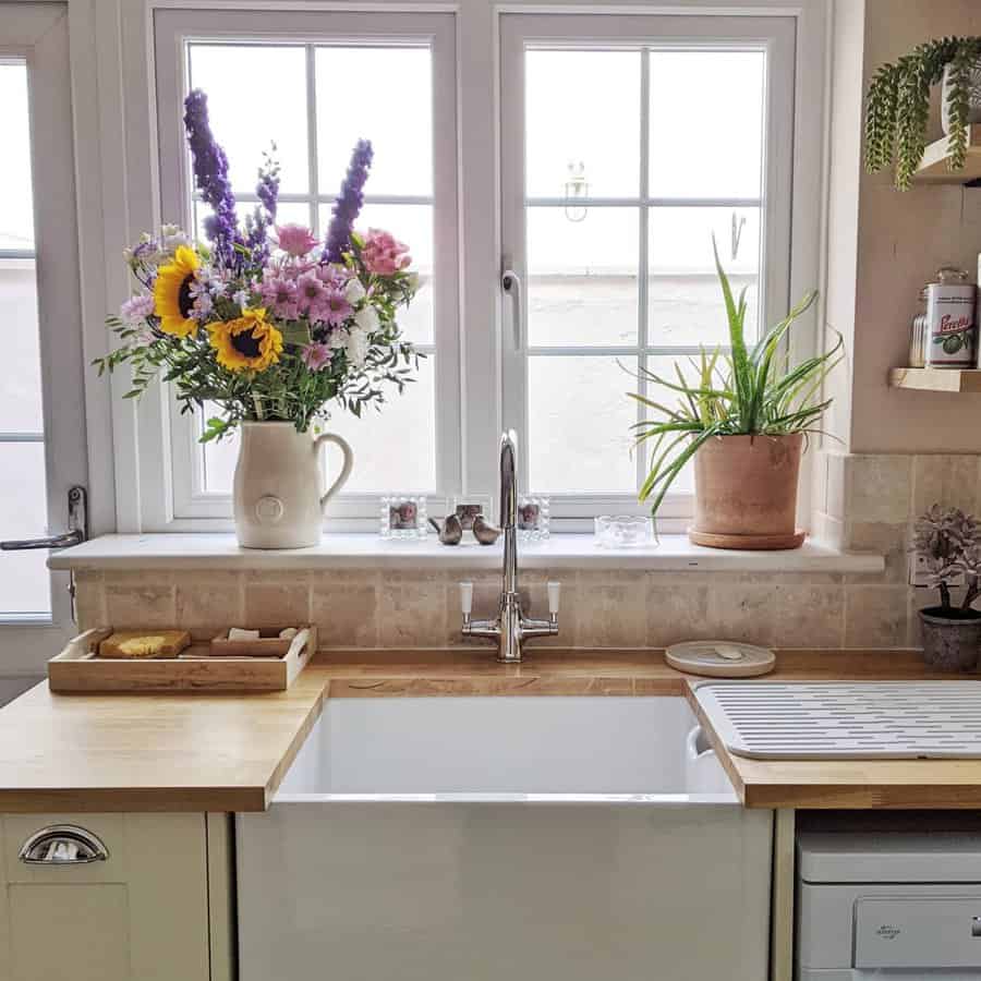 A single kitchen sink with wooden countertops, sunflowers in a jug, a potted plant, and a drying rack beneath a window