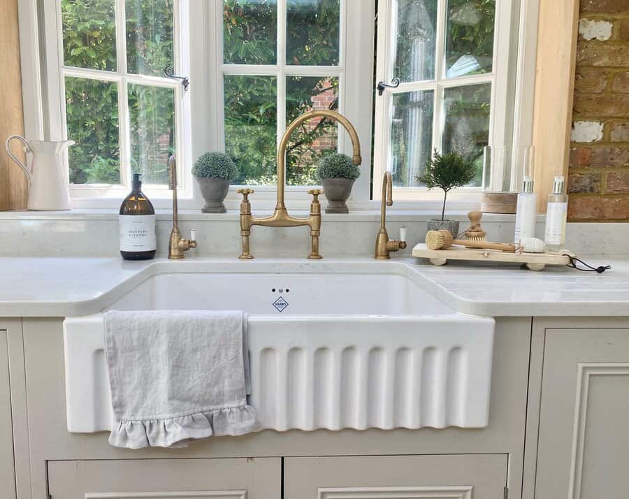 A farmhouse kitchen sink with a brass faucet, surrounded by potted plants, soap, and a towel, in front of a window with garden view