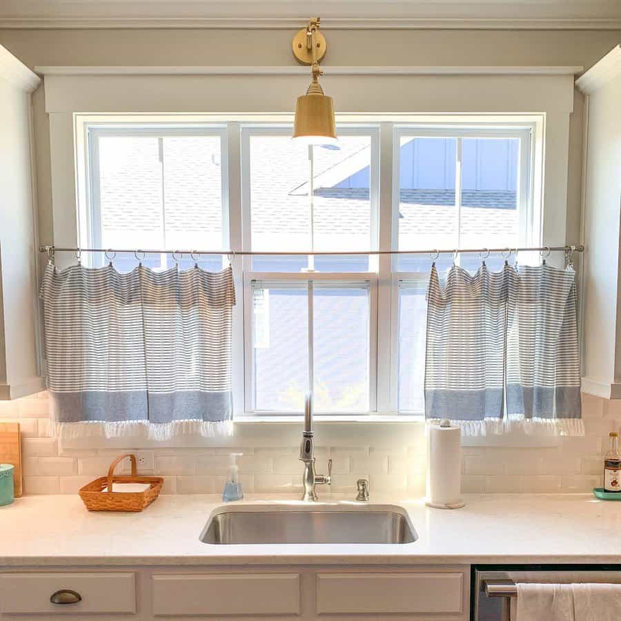 Kitchen sink with striped curtains and a gold wall lamp above, bright natural light from the window, wicker basket on the counter