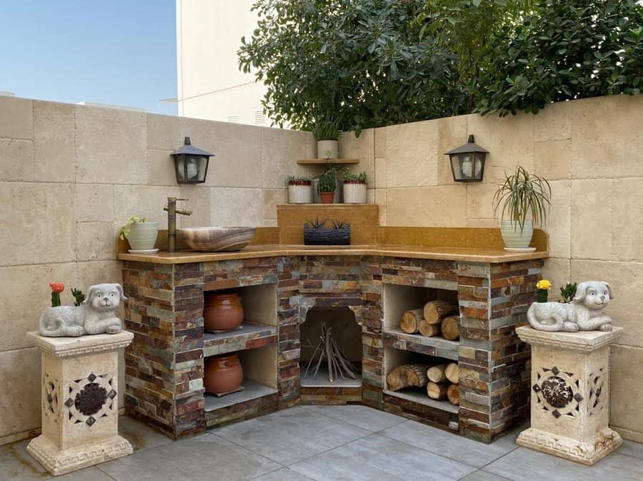 Outdoor corner stone fireplace with wood storage, decorative pots, plants, and lanterns, flanked by dog statues on pedestals