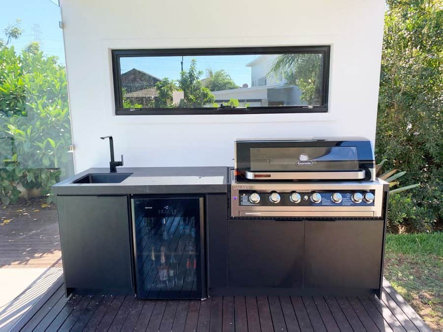 Outdoor kitchen with a sleek black countertop, sink, mini-fridge, and grill set on a wooden deck, surrounded by greenery