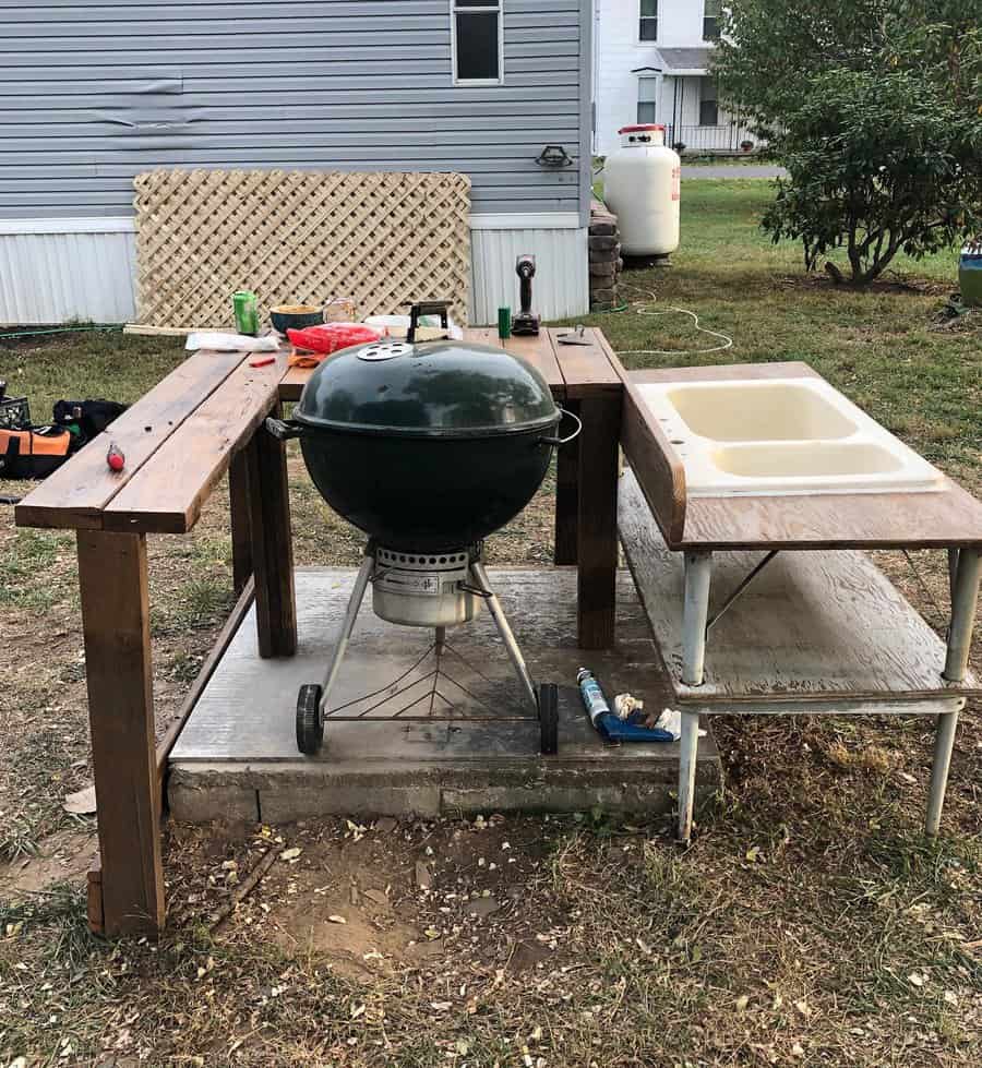 Outdoor cooking station with a green charcoal grill, wooden table, and sink on a concrete slab beside a house, tools are on the table