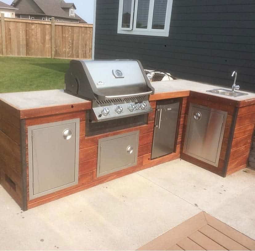 Outdoor kitchen setup with a stainless steel grill, sink, cabinets, and fridge on a concrete patio next to a fenced lawn