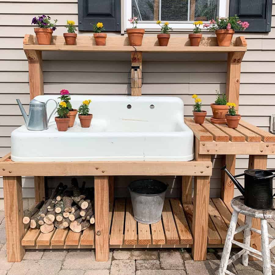 Outdoor potting bench with a white sink, potted flowers, a watering can, and firewood storage underneath against a house exterior