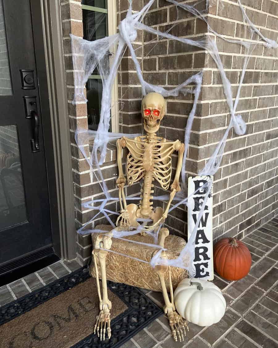 Skeleton with red eyes on porch, surrounded by spider webs and pumpkins; "Beware" sign nearby