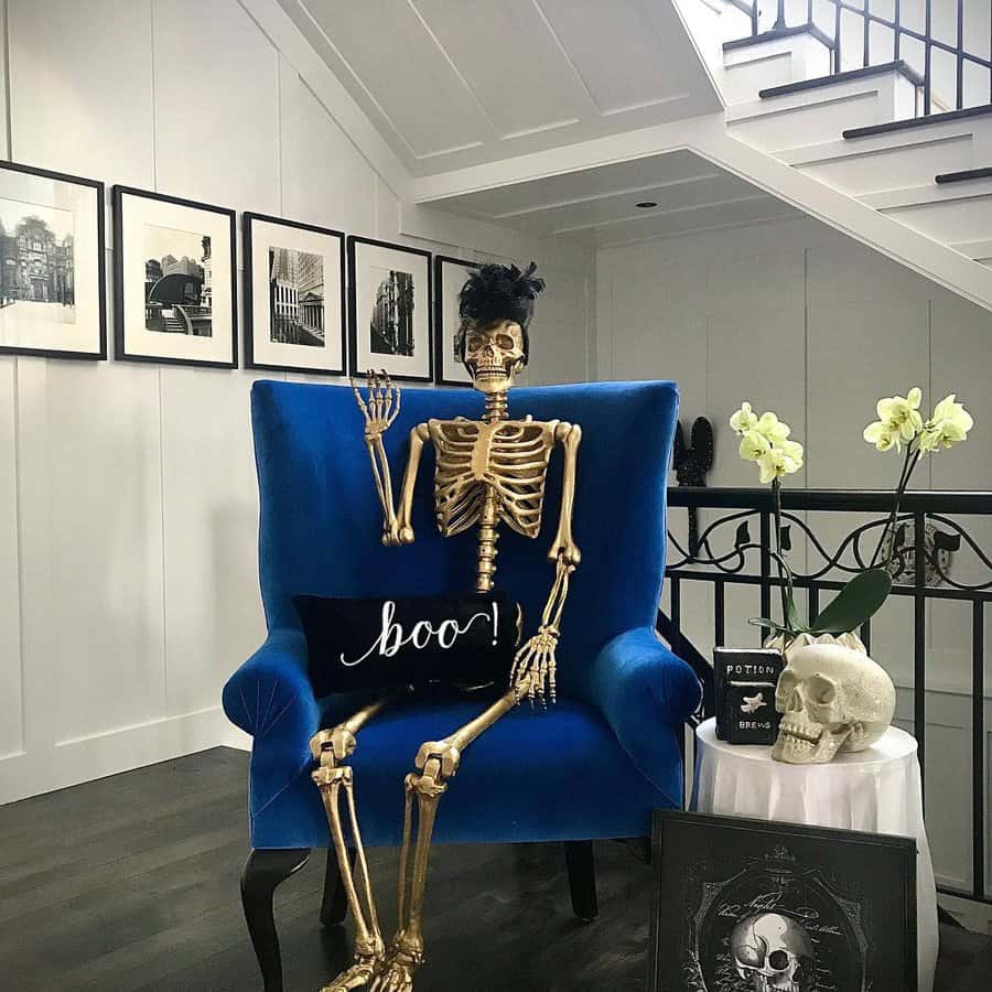 Skeleton in a blue armchair with a "boo!" pillow, Halloween decor, and skulls nearby