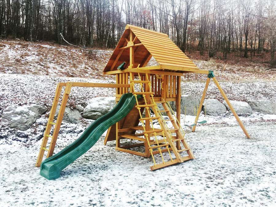 Wooden playground set with slide, swings, and climbing ladder, on snow-dusted ground, and trees in the background