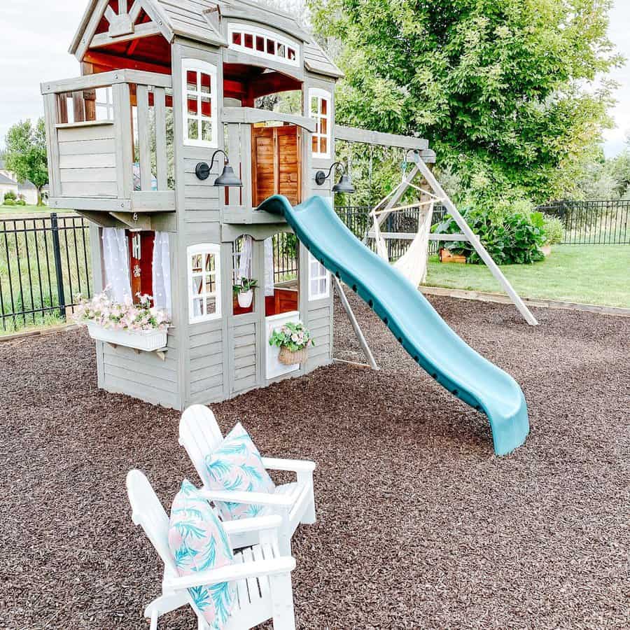 Playhouse with a blue slide, swings, and white chairs with pillows on a mulched surface, surrounded by greenery and a black fence