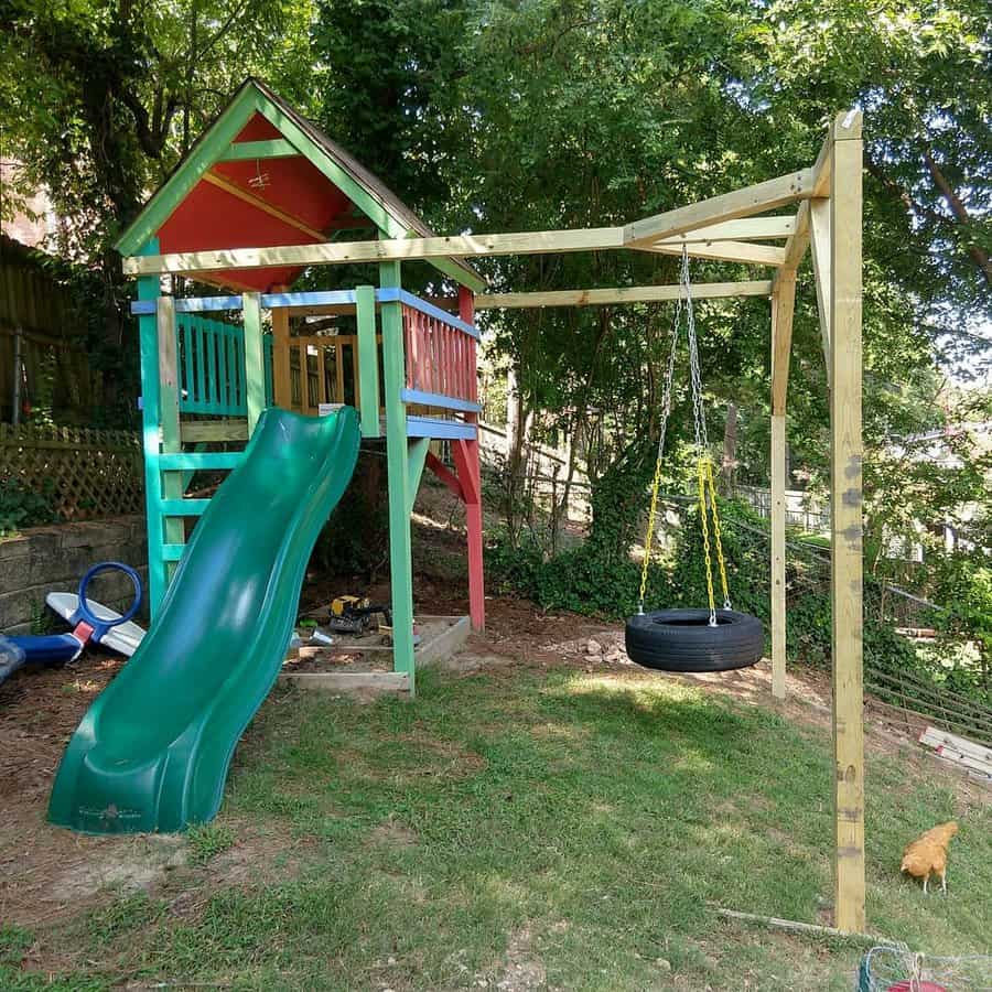 Wooden playset with green slide, tire swing, and colorful playhouse in a grassy backyard surrounded by trees and a fence