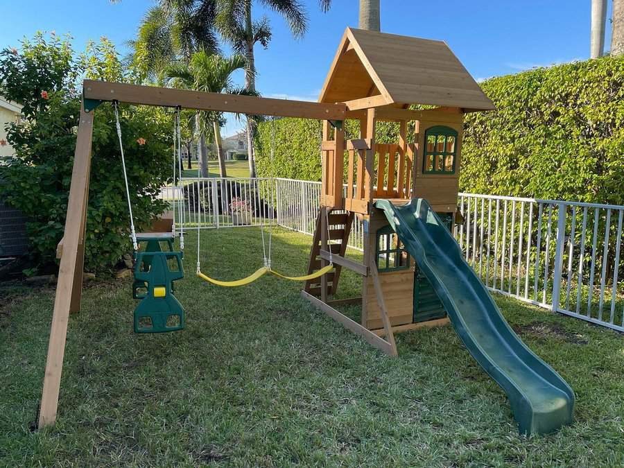 Wooden playset with slides, swings, and a climbing area in a sunny, fenced backyard with palm trees and hedges