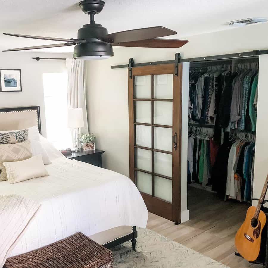 Bedroom with a bed, white bedding, wooden sliding closet door, ceiling fan, nightstand with lamp, and a guitar leaning on the wall