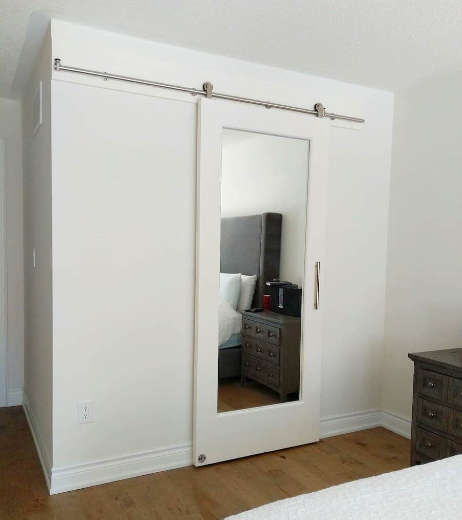 Sliding barn door with a full-length mirror in a bedroom, adjacent to a bed and dresser on wooden floor