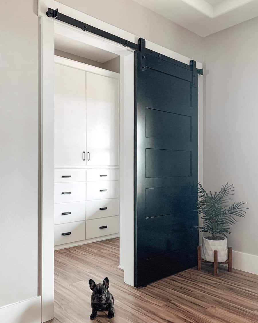 A sleek black barn-style sliding door with black hardware adds contrast to a modern closet space with white cabinetry and wood flooring