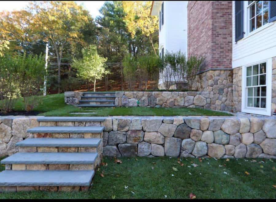 Tiered stone retaining walls with built-in steps, landscaped greenery, and a sloped yard adjacent to a brick and white house