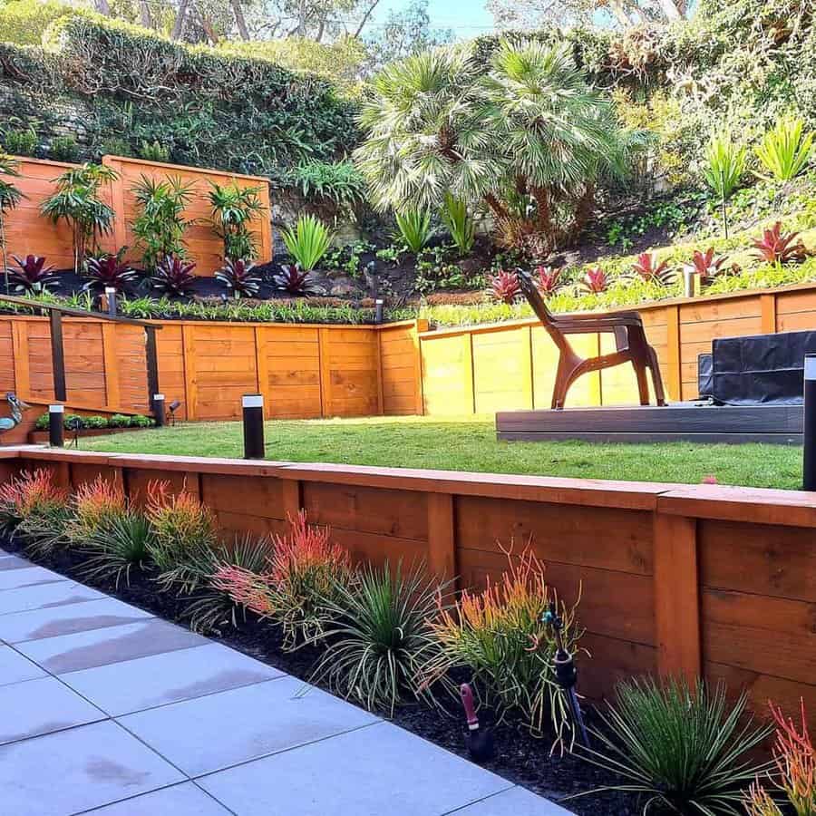 Multi-level wooden retaining walls with vibrant landscaping, tropical plants, a lush green lawn, and a patio area with pavers